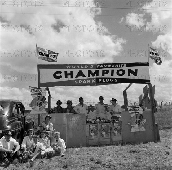 Champion spark plugs. A European family enjoy a picnic in front of the Champion stand at the Aquasports Regatta. A sign above the stand reads: WORLD'S FAVOURITE CHAMPION SPARK PLUGS. Kenya, 28 October 1956. Kenya, Eastern Africa, Africa.