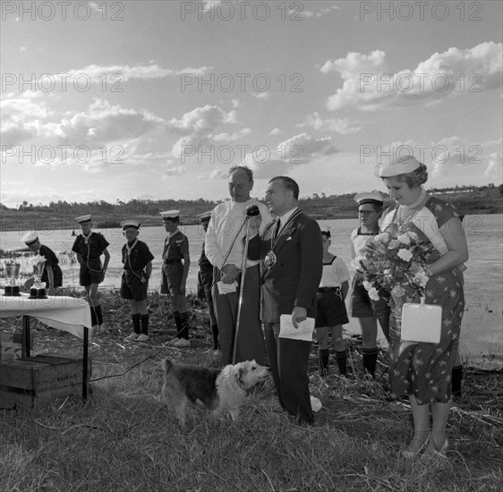 Prize-giving ceremony at the Aquasports Regatta. A small dog takes centre stage as a suited man wearing a large medal talks into a microphone at the prize-giving ceremony of the Aquasports Regatta. Boy scouts in naval uniform stand in line behind him. Kenya, 28 October 1956. Kenya, Eastern Africa, Africa.
