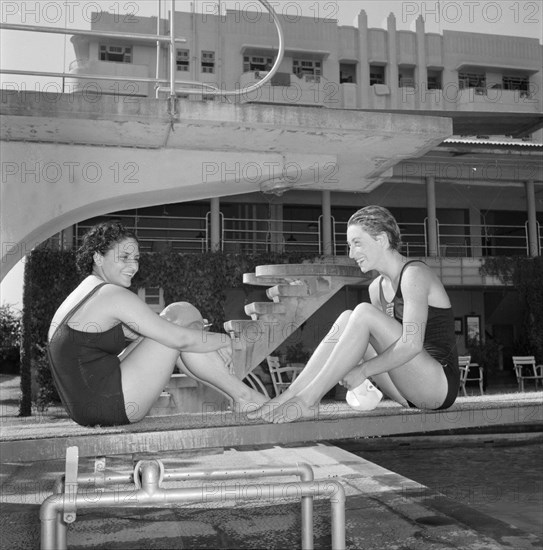 Facing the opposition. Two female swimmers competing in the 1956 Olympic Games sit facing each other wearing swimming costumes on a diving board. Kenya, 2 November 1956. Kenya, Eastern Africa, Africa.