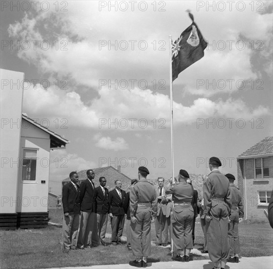 The 1956 Ugandan Olympic team. Athletes from the 1956 Ugandan Olympic team stand in line with officials as their national flag is raised in a Ugandan village. Uganda, 5 November 1956. Melbourne, Victoria, Australia, Australia, Oceania.