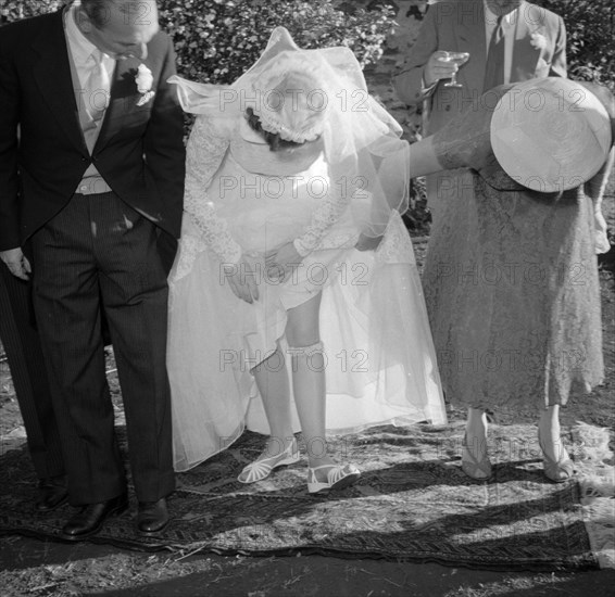 A bride reveals her garter. The bride at the Carey-Courtney wedding lifts up her wedding dress to reveal her garter. Kenya, 15 December 1956. Kenya, Eastern Africa, Africa.