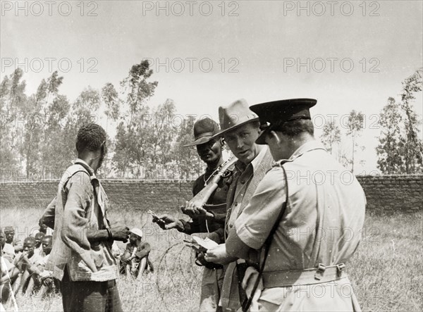 Searching for Mau Mau suspects. A farmer and military officers question a Kikuyu farm labourer. Behind him more Kikuyu sit on the ground surrounded by coils of barbed wire. Kenya, circa 1953. Kenya, Eastern Africa, Africa.