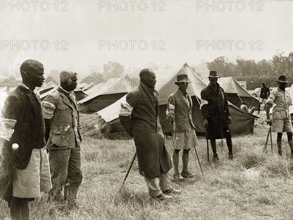 Kikuyu Home Guards in camp. Six Kikuyu Home Guards, dressed in makeshift uniforms and armed with spears, stand in front of rows of substantial tents, presumably a temporary home guard post. Kenya, circa 1952. Kenya, Eastern Africa, Africa.