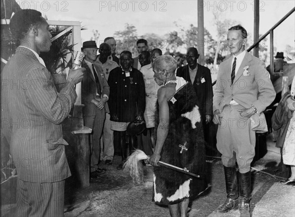 Maina arap Chumo at a function. Maina arap Chumo, a Kikuyu elder dressed in a cowhide wrap and carrying a whisk, arrives or speaks at an official or settler gathering in Nakuru. A radio interviewer stands nearby. Nakuru, Kenya circa 1953. Nakuru, Rift Valley, Kenya, Eastern Africa, Africa.