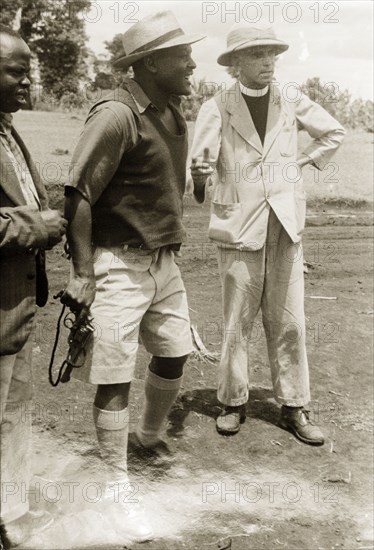 Pro-government Kikuyu and a Catholic priest. A loyal Kikuyu chief talks to a Roman Catholic priest from a nearby Mission as he hunts gangsters, revolver in hand. Kenya, circa 1953. Kenya, Eastern Africa, Africa.