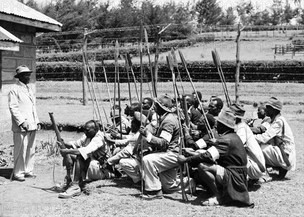 Chief Muhoya addresses the Home Guard. An official photograph of Chief Muhoya of Nyeri speaking to a group of Kikuyu Home Guards who squat on the ground within a barbed-wire compound. Most are armed with spears, but the foremost guard carries a rifle. Nyeri, Kenya, 1952. Nyeri, Central (Kenya), Kenya, Eastern Africa, Africa.