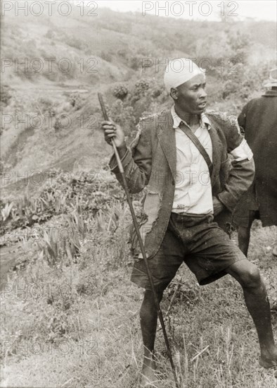 A Kikuyu Home Guard on duty. A young man in a tattered jacket stands barefoot on a hillside, holding a spear. A white cloth around his arm and head indicates he is a member of the Kikuyu Home Guard. Kenya, circa 1952. Kenya, Eastern Africa, Africa.