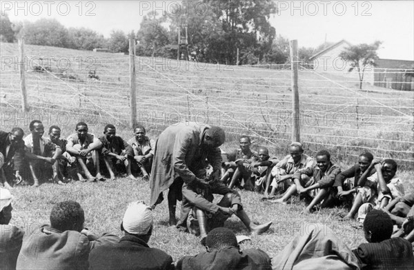 Government de-oathing ceremony. A Mau Mau detainee sitting in the centre of a circle of men in a fenced compound is 'purified' of the Mau Mau oath. Kenya, circa 1954. Kenya, Eastern Africa, Africa.