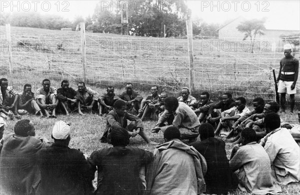 Government de-oathing ceremony. A Mau Mau detainee sitting in the centre of a circle of men in a fenced compound is prompted to confess to having taken the Mau Mau oath. Kenya, circa 1954. Kenya, Eastern Africa, Africa.