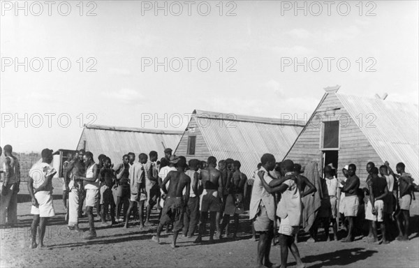 Home Guards at their living quarters. Young Kikuyu men gather beside prefabricated huts, a temporary accommodation camp for the Kikuyu Home Guard. Kenya, circa 1953. Kenya, Eastern Africa, Africa.