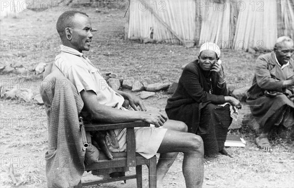Kikuyu screening team. A Kikuyu Home Guard is seated in a chair beside two Kikuyu men who have been interviewing Mau Mau suspects. Kenya, circa 1954. Kenya, Eastern Africa, Africa.