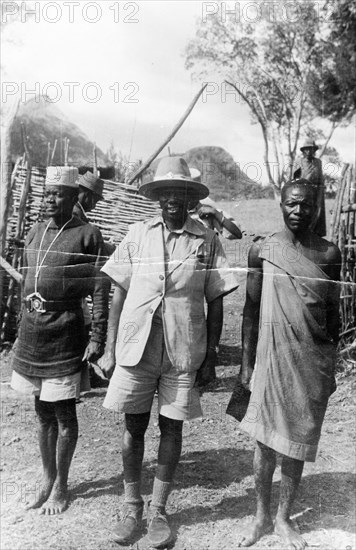Mau Mau suspect and guards. A Mau Mau suspect under arrest is guarded by two men who stand either side of him, one wearing military uniform, the other wearing a traditional cloth wrap. Kenya, circa 1953. Kenya, Eastern Africa, Africa.