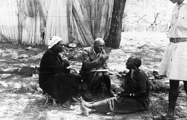 Interviewing a Mau Mau suspect. Two Kikuyu men gesticulate at an older Kikuyu woman seated on the ground whom they believe has taken two Mau Mau oaths. Kenya, circa 1954. Kenya, Eastern Africa, Africa.