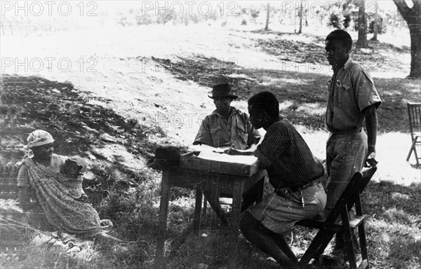 Questioning a Mau Mau suspect. Three men in lightweight uniforms question a Kikuyu woman with a baby whom they believe may be a Mau Mau suspect. Kenya, circa 1954. Kenya, Eastern Africa, Africa.