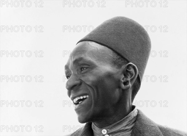 Kikuyu man. Portrait of a smiling Kikuyu man in shirt, jacket and fez-like hat. The photograph does not reveal his identity. Kenya, circa 1953. Kenya, Eastern Africa, Africa.