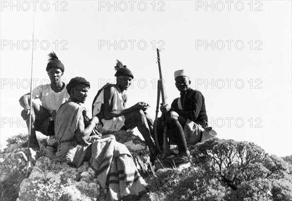 Kikuyu Home Guards on patrol. Four Kikuyu Home Guards on patrol sit atop a rocky outcrop. One wears a uniform and carries a rifle, another holds a spear. Kenya, circa 1952. Kenya, Eastern Africa, Africa.