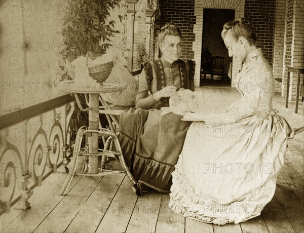 Hobbies on the veranda. Portrait of Ellen May Brodribb doing needlework on the veranda of a colonial house. She is accompanied by an unidentified woman whose head is bowed, intent on reading a book. Near Brisbane, Australia, circa 1895., Queensland, Australia, Australia, Oceania.