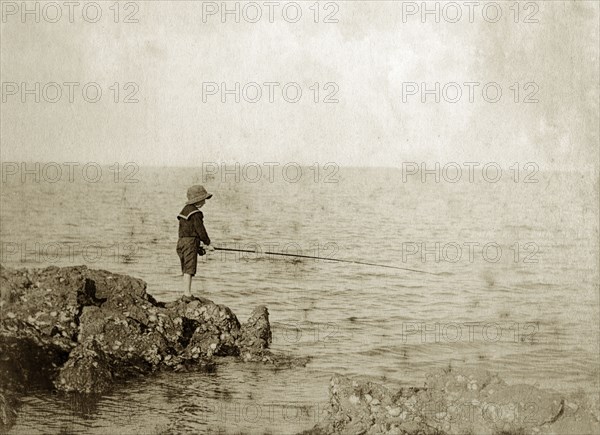 Young boy fishing, Australia. A lone boy wearing a sailor suit and hat, holds his fishing line out over rocks into the sea in the hope of catching a bite. Queensland, Australia, circa 1890., Queensland, Australia, Australia, Oceania.