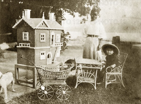 Marjory Wood's dollhouse. A young girl identified as 'Marjory Wood' sits at a miniature table beside a large dollhouse wearing an outsized solatopi hat. Her Jarawan ayah (nursemaid) stands over her, dressed in traditionally Western-style clothing. Andaman Islands, India, circa 1908., Andaman and Nicobar Islands, India, Southern Asia, Asia.