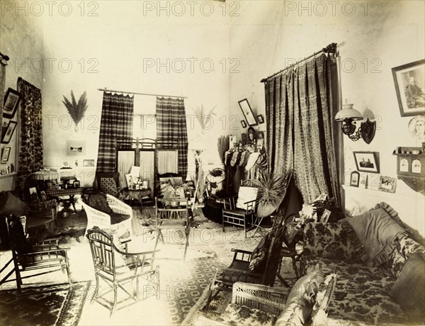 Mrs Morton's drawing room. Interior shot of a traditionally Victorian drawing room belonging to 'Mrs Morton'. Characterised by a high ceiling and floor-length windows, the room is busily decorated with furniture, framed photographs and heavily patterned fabrics. India, circa 1895. India, Southern Asia, Asia.