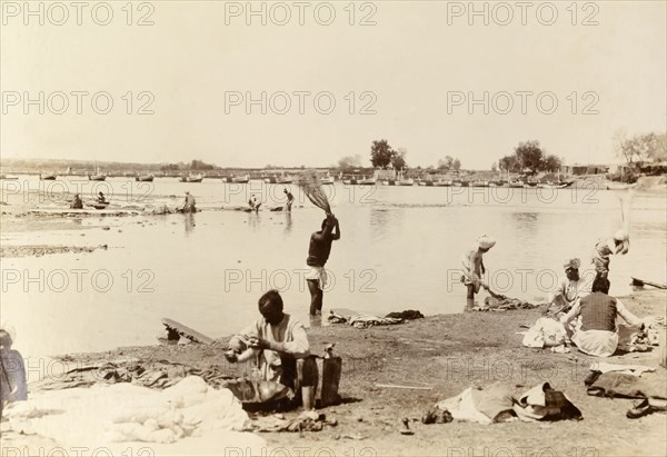 Washing clothes in the river. Indian men wash items of clothing by thrashing them onto special stones at the edge of a wide river. India, circa 1895. India, Southern Asia, Asia.