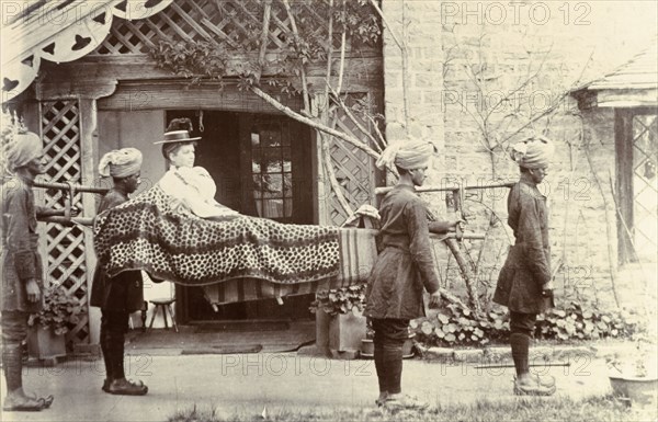 Mem Sahib Blount goes visiting. Mem Sahib Blount reclines in an open sedan chair as she is carried by a group of Indian servants dressed in turbans and curly-toed shoes. India, circa 1895. India, Southern Asia, Asia.