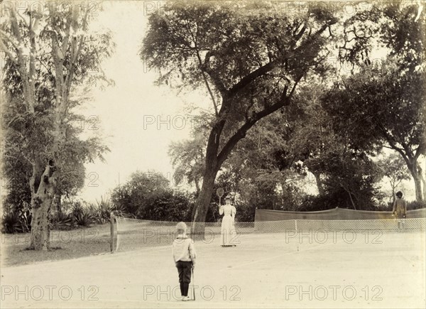 Mrs Page plays tennis. A British women identified as 'Mrs Page' holds up her tennis racquet towards her opponent, a small boy who faces her across the net of a large tennis court. Dressed in sporting whites, her look is completed with a stylish straw boater. India, circa 1895. India, Southern Asia, Asia.