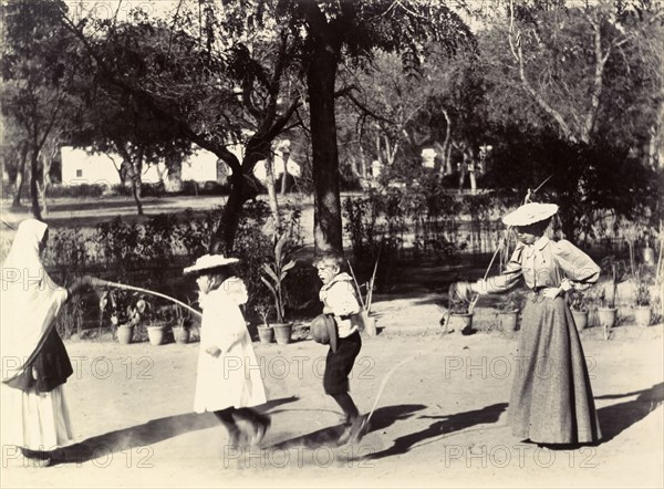 Skipping in India. Two women, one British and one Indian, swing a skipping rope in a garden for two British children who jump over it with delight. India, circa 1895. India, Southern Asia, Asia.