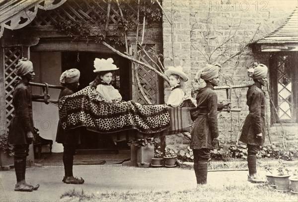 A four-anna ride'. Two British girls dressed in their best hats sit opposite each other in an open sedan chair carried by a group of Indian servants dressed in turbans and curly-toed shoes. India, circa 1895. India, Southern Asia, Asia.