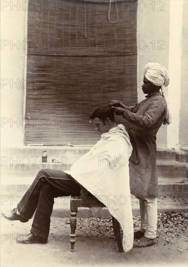 Cut your hair Sir'. A turbaned Indian barber trims the hair of a British gentleman who sits patiently, draped in a towel with his head bowed. India, circa 1895. India, Southern Asia, Asia.