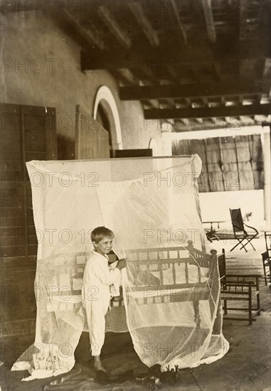 Mosquito curtains. A young boy dressed in pyjamas stands, resting one foot on a bed shrouded with mosquito curtains. India, circa 1895. India, Southern Asia, Asia.