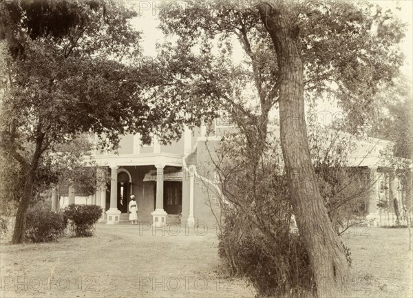 Officer's bungalow, Pindi. View through the trees of a uniformed Indian servant at the columned entrance to an officer's bungalow. Rawalpindi, Punjab, India (Punjab, Pakistan), circa 1895. Rawalpindi, Punjab, Pakistan, Southern Asia, Asia.