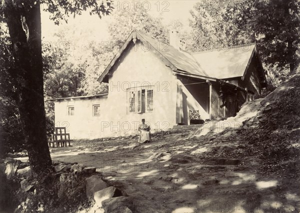Mrs Page reads in the garden. A distant figure identified as 'Mrs Page' sits reading her book in front of a bungalow. The original caption suggests she is 'watching tongas' (two-wheeled carriages drawn by horses). India, circa 1890. India, Southern Asia, Asia.