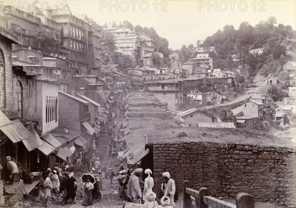 Bustling market street at Murree. Shop awnings line a bustling market street that winds down from the hills into the town of Murree. A lone British women holding a parasol can be see amongst the crowds of Pakistani locals. Murree, Punjab, India (Punjab, Pakistan), circa 1895. Murree, Punjab, Pakistan, Southern Asia, Asia.