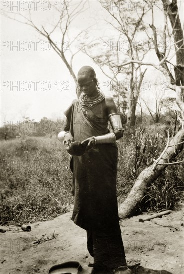 A Maasai woman. A Maasai woman in traditional dress and jewellery including ornate necklaces and armbands, examines the contents of a small bowl. The original caption identifies her as a 'demure wa woman'. Probably Tanganyika Territory (Tanzania), circa 1930. Tanzania, Eastern Africa, Africa.