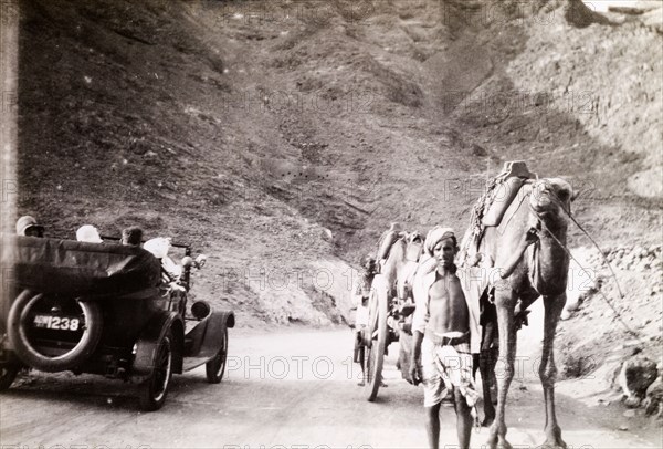 Road traffic near Suez. An Egyptian man leads a camel and cart along a winding road past a motorcar full of Europeans travelling in the other direction. Suez, Egypt, circa 1930. Suez, Suez, Egypt, Northern Africa, Africa.