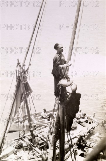 A 'gully gully' man. An Egyptian 'gully gully man' (travelling magician), perches high on the mast of a small sailing vessel, rope and basket in hand. Suez, Egypt, circa 1930. Suez, Suez, Egypt, Northern Africa, Africa.