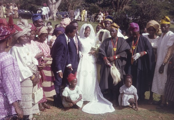Sayo Bewaji's wedding'. Originally captioned as 'Sayo Bewaji's wedding', a bride and groom pose for the camera surrounded by family wedding guests who wear traditional Nigerian dress. Nigeria, circa 1972. Nigeria, Western Africa, Africa.