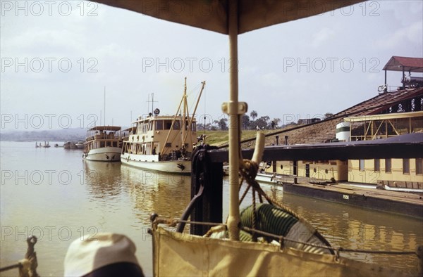 Lokoja trading houses and ferries. Two large ferries docked on the riverside near the Royal Niger Company trading houses. Lokoja, Nigeria, circa 1965. Lokoja, Kogi, Nigeria, Western Africa, Africa.
