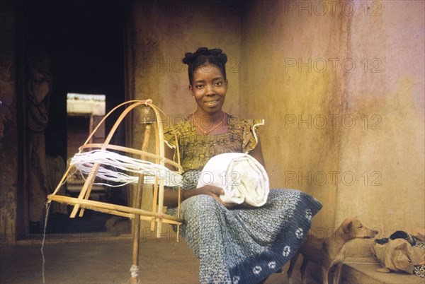 Spinning thread, Nigeria. A young woman sits beside a homemade spinning frame, a bundle of cloth resting on her knee. Okene, Nigeria, circa 1964. Okene, Kogi, Nigeria, Western Africa, Africa.