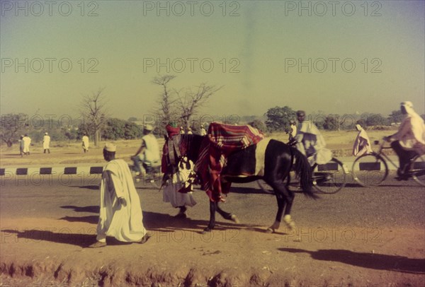 Hurrying to a celebration. Pedestrians and cyclists hurry along a road to a celebration marking the end of Ramdan. The horse pictured has had its bridle decorated to participate in a religious procession. Zaria, Nigeria, 1958. Zaria, Kaduna, Nigeria, Western Africa, Africa.