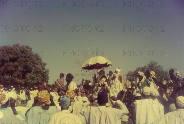 The Emir of Zaria. The 'Sarkin Zazzau' (Emir of Zaria), Alhaji Shehu Idris, is protected from the sun by a tassled parasol as he rides horseback in a procession celebrating the end of Ramadan. Zaria, Nigeria, 1958. Zaria, Kaduna, Nigeria, Western Africa, Africa.