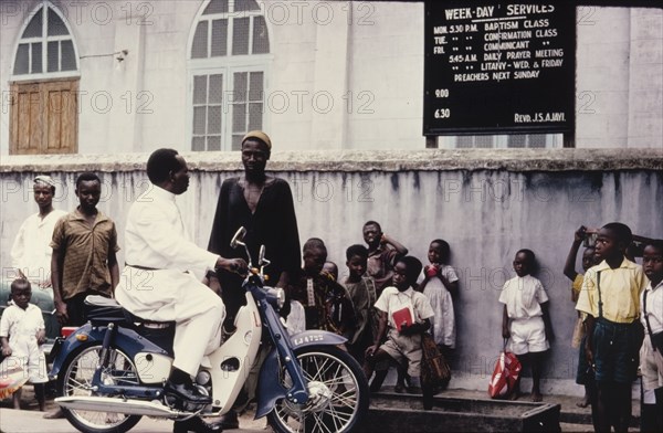 Pastor on a moped. Reverend J. Harwood, a Church of Nigeria pastor, pulls over his moped to chat with a man outside a church. A group of children stand leaning against the church wall. Nigeria, circa 1968. Nigeria, Western Africa, Africa.