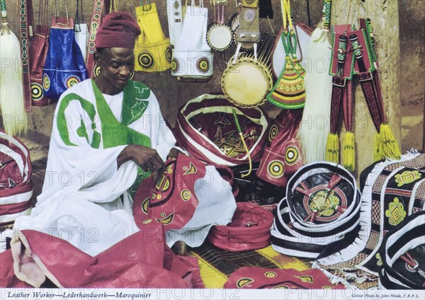 A leather craftsman, Nigeria. A colourful tourist postcard depicts a Nigerian leather craftsman at work, sitting cross-legged on the ground as he embroiders a leather bag. A selection of completed bags and other leather goods surround him. Nigeria, circa 1975. Nigeria, Western Africa, Africa.