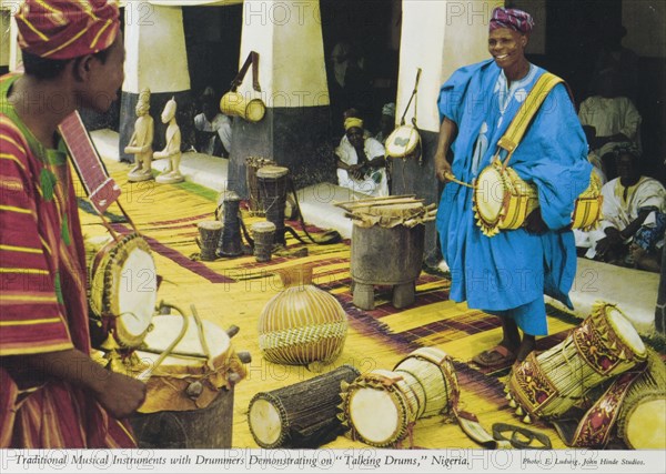 Talking drums', Nigeria. A colourful tourist postcard depicts two Nigerian drummers playing on 'talking drums' before a crowd of onlookers. Surrounded by traditional drums of all shapes and sizes, they perform on rush mats laid down on the street. Nigeria, circa 1975. Kano, Kano, Nigeria, Western Africa, Africa.