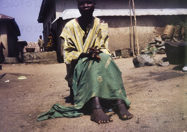 Psychiatry patient at Aro. Portrait of a female psychiatry patient, pictured outdoors at Aro Mental Hospital. An original caption suggests she has been 'restrained from self-harm', apparent from the metal shackles fitted around her ankles. Abeokuta, Nigeria, circa 1971. Abeokuta, Ogun, Nigeria, Western Africa, Africa.