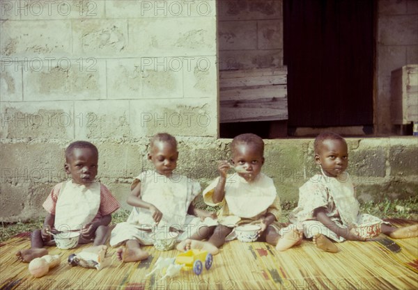 Orphans at a Church of Nigeria hospital. Four young orphans, seated on a rush mat, feed themsleves with bowls and spoons outside a Church of Nigeria hospital. Nigeria, circa 1962. Nigeria, Western Africa, Africa.