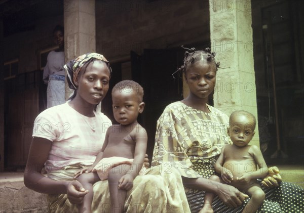 Patients at a Church of Nigeria Hospital. Portrait of two Nigerian women, seated outside a Church of Nigeria hospital with their young children on their laps. An original caption identifies them as hospital patients. Nigeria, circa 1970. Nigeria, Western Africa, Africa.