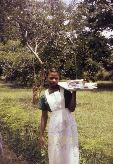 Training midwife, Nigeria. Outdoors portrait of a young woman training to become a midwife for a Church of Nigeria hospital. Dressed in uniform, she balances a tray of medical equipment on one hand. Nigeria, circa 1970. Nigeria, Western Africa, Africa.