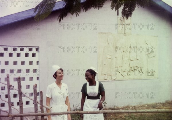 Nurses at a Church of Nigeria hospital. A European nurse and an African midwife share a joke outside the children's ward of a Church of Nigeria hospital. Nigeria, circa 1970. Nigeria, Western Africa, Africa.